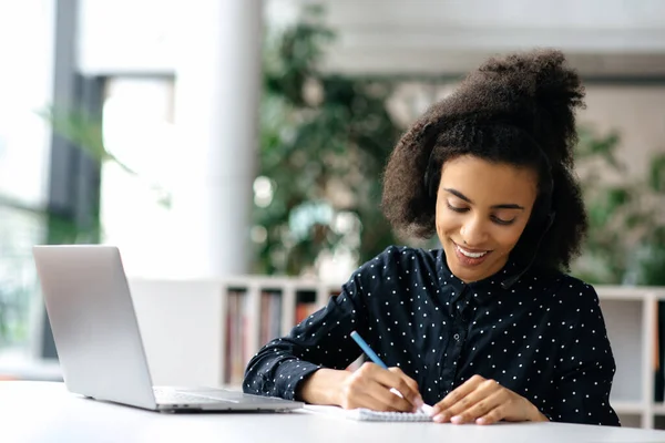 Jeune femme afro-américaine focalisée avec casque, vêtue d'une chemise formelle, manager ou pigiste, assise à table au bureau, travaillant sur un ordinateur portable, prenant des notes, souriant — Photo