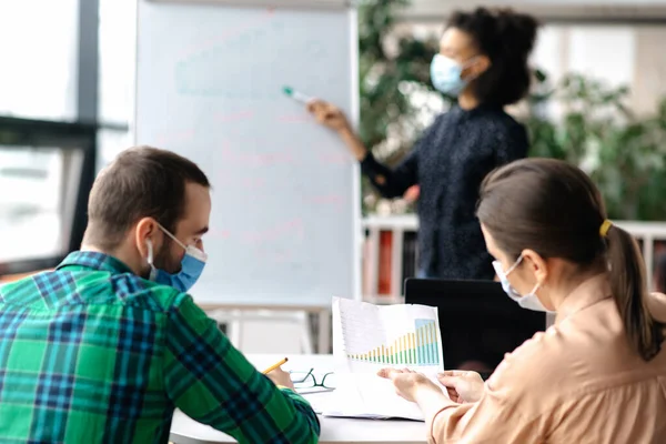 Empleados exitosos se reunieron en la oficina con máscaras médicas protectoras, discutiendo un proyecto empresarial, contra el fondo es borrosa mujer afroamericana que muestra información en pizarra blanca — Foto de Stock