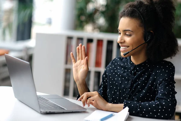 Wanita muda african Amerika yang gembira dengan headset, manajer sukses atau operator pusat panggilan, duduk di kantor, menggunakan laptop, berbicara pada konferensi video dengan klien atau karyawan, melambaikan tangan — Stok Foto