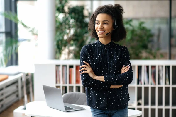 Portrait d'une jolie, agréable jeune femme afro-américaine, avec casque et dans l'usure élégante, freelance, travailleur de centre d'appels ou opérateur de soutien, se tient près du lieu de travail, regarde loin, bras croisés, sourire — Photo