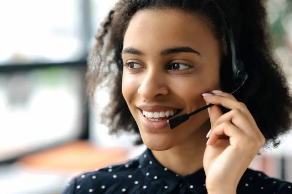 Gros plan de charmante jeune femme afro-américaine aux cheveux bouclés en casque et chemise stylée, travailleuse du centre d'appels ou opératrice de soutien, regardant vers le côté, souriante et amicale — Photo