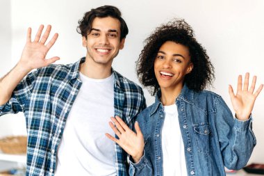 Kameraya poz veren neşeli adam ve kız. Dost canlısı, neşeli, çok ırklı genç bir çiftin portresi günlük şık kıyafetler içinde, el hareketleriyle webcam 'da karşılanıyor, kameraya bakın, mutlu bir şekilde gülümsüyor.