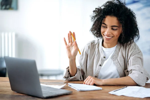 Reunión de video en línea. Chica afroamericana alegre, estudiante o freelancer, comunicarse en videoconferencia con compañeros de trabajo o amigos utilizando el ordenador portátil, saludo con gesto de mano, sonriendo amigable —  Fotos de Stock