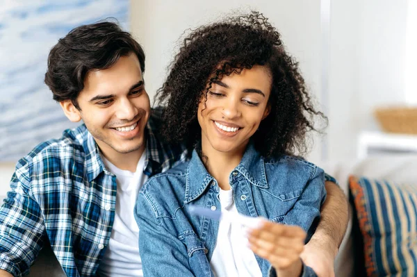 Joven feliz mixta raza casada pareja sentado en sofá mirando positivo embarazo prueba disfrutando de buenas noticias, preparándose para convertirse en padres, africano americana chica tiene positivo embarazo prueba en mano — Foto de Stock