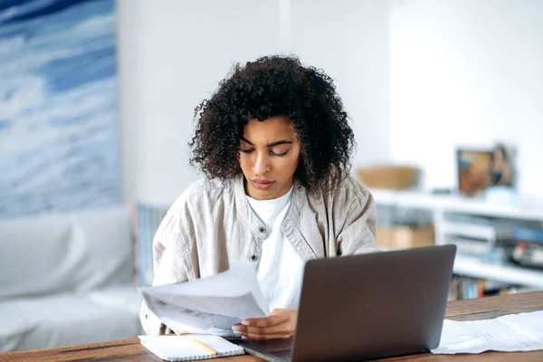 똑똑 한 집중력의 곱슬머리의 아프리카 계 미국인 여성 평상복을 입고 프리랜서나 학생 이 거실에 앉아 노트북 컴퓨터로 일하거나 원격으로 공부하거나 서류를 보면서 — 스톡 사진
