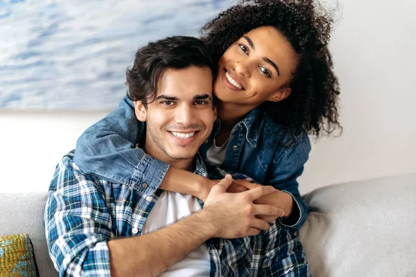 Retrato alegre feliz pareja de raza mixta, chica rizada bastante afroamericana abrazando tiernamente a su novio hispano sentado en el sofá, usando ropa casual en la sala de estar, mirando a la cámara, sonriendo — Foto de Stock