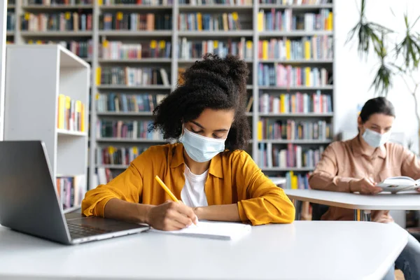 Estudantes multiétnicos estudam durante a pandemia, sentam-se em mesas à distância em máscaras médicas protetoras na biblioteca com um laptop, ouvem palestras, menina afro-americana toma notas — Fotografia de Stock