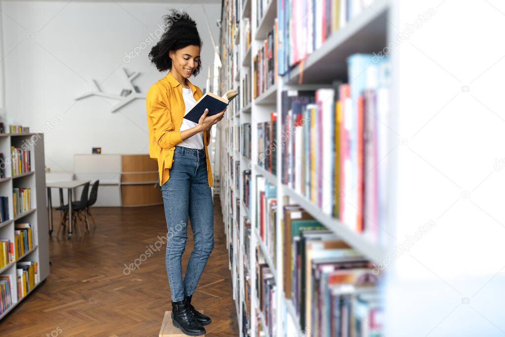 Happy joyful smart african american curly haired female student in casual stylish clothes, standing near the bookshelf in university library, is choosing a book, reading while standing, smiling