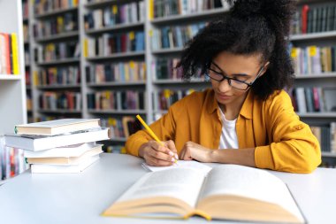 Başarılı zeki genç kadın, gözlüklü Afro-Amerikan kız öğrenci, üniversite kütüphanesinde masada oturuyor, derse ya da sınava hazırlanıyor, ödevlerini yapıyor, not defterine notlar yazıyor.