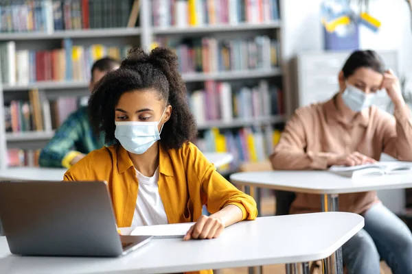 Jovens estudantes estudam durante a pandemia, sentados na universidade à distância uns dos outros usando máscaras médicas protetoras. Estudante afro-americana tomando notas durante palestra, usando laptop — Fotografia de Stock