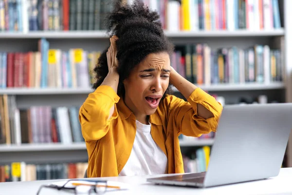 Kesal gadis, Afrika Amerika mahasiswa perempuan, duduk di meja di perpustakaan universitas, mencengkeram kepalanya dalam horor, melihat layar laptop, merasa stres, mendapat berita negatif — Stok Foto