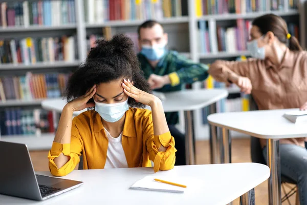 Estudantes multirraciais estudam durante uma pandemia, sentam-se em máscaras médicas protetoras à distância um do outro, meninas afro-americanas melodias para a lição, contra os estudantes de fundo são cumprimentar seus cotovelos — Fotografia de Stock