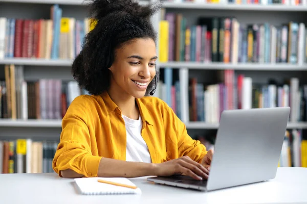 Heureuse jeune fille afro-américaine séduisante en vêtements élégants, étudiante assise dans la bibliothèque de l'université au bureau, se préparant pour l'examen ou la leçon, naviguant sur Internet, cherchant des informations, souriant — Photo