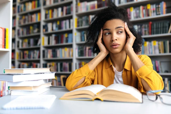 疲惫不堪的非洲裔美国女学生，坐在校园图书馆的桌旁，休息一下看书，准备上课或考试，回头看，头疼，需要休息 — 图库照片