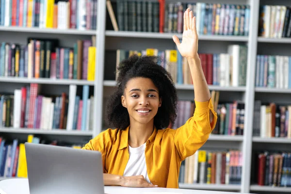 Excellent female student. Smart african american student girl at a lecture sits at a table, stylishly dressed, raises her hand, knows the answer to question and has an idea, wants to answer, smiles