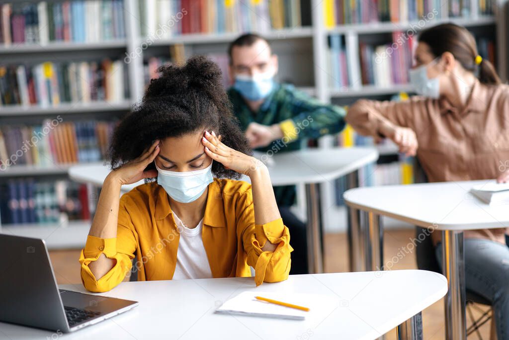 Multiracial students study during a pandemic, sit in protective medical masks at distance from each other, African-American girl tunes to lesson, against the background students are greet their elbows