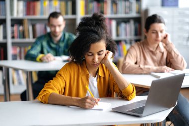 Dersteki öğrenciler. Üniversite kütüphanesinde masada oturan, derslere ya da sınavlara hazırlanan, notlar alan, arka plandaki odaklı olmayan öğrencileri düşünen, konsantre olmuş ciddi Afrikalı Amerikalı kız öğrenci.
