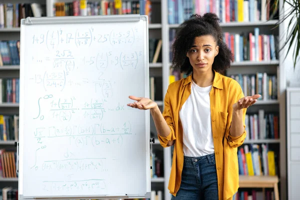 Porträt einer verwirrten jungen afrikanisch-amerikanischen Frau, die neben Whiteboard steht, lässig gekleidet ist, Unterricht leitet, mit Studenten online kommuniziert und überrascht in die Kamera blickt — Stockfoto