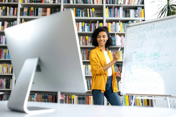 Радісна афроамериканська жінка-тренер, що стоїть біля дошки, в повсякденному одязі, проводить онлайн-вебінар, спілкується зі студентами за викликом конференції, дивлячись на екран комп'ютера, посміхаючись — стокове фото