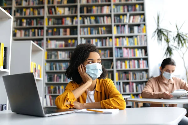 Duas estudantes inteligentes de diferentes nacionalidades em máscaras médicas estão sentadas em mesas na biblioteca da universidade, preparando-se para aulas ou um exame, uma menina afro-americana olha pensativamente para longe — Fotografia de Stock