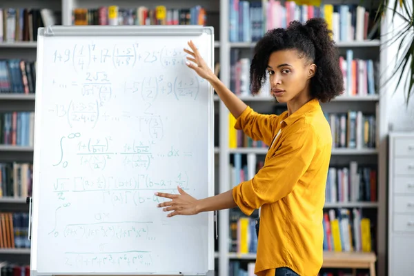 Ernste junge Frau, gemischte Rassenlehrerin, die neben Whiteboard steht, lässig gekleidet, erklärt Thema für Schüler per Videokonferenz, gestikuliert mit den Händen, blickt in die Kamera — Stockfoto