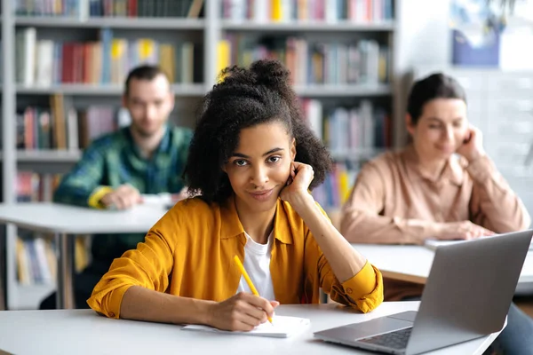 Studenten in der Vorlesung. Konzentrierte afrikanisch-amerikanische Studentin sitzt am Tisch in der Universitätsbibliothek, bereitet sich auf Unterricht oder Prüfung vor, blickt in die Kamera, defokussierte Studenten im Hintergrund — Stockfoto