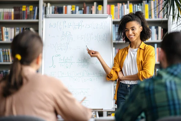 Successo piacevole afro-americano ricci capelli giovane insegnante donna in piedi presso la lavagna in biblioteca, dando una lezione per gli studenti, mostra informazioni e sorridente — Foto Stock