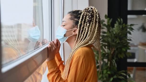 African American girl in a medical protective mask at home in quarantine, went to the window, looked sadly out the window at the street, she is at home on self-isolation due to illness — Stock Video