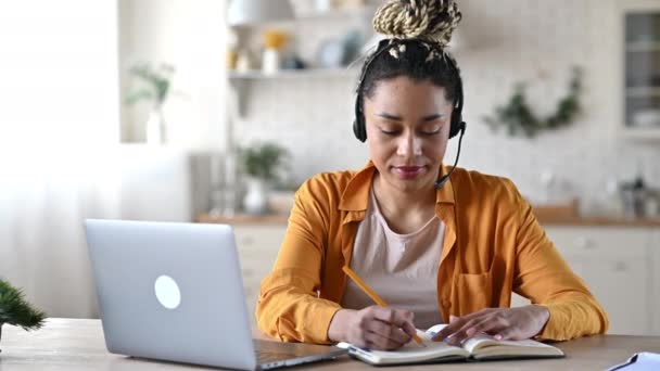 Amistosa joven atractiva mujer afroamericana en auriculares, empleado del centro de llamadas, operador de soporte, trabajando en una computadora portátil, lleva a cabo una consulta en línea con el cliente, tomando notas, sonriendo — Vídeos de Stock