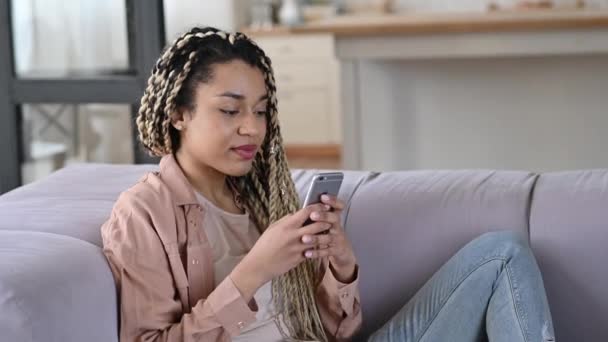 Satisfeito alegre menina afro-americana senta-se em um sofá, usando smartphone, passar o tempo nas mídias sociais, tem uma boa notícia ou mensagem, navegar na internet, conversando on-line com amigos ou familiares, ganhar loteria — Vídeo de Stock