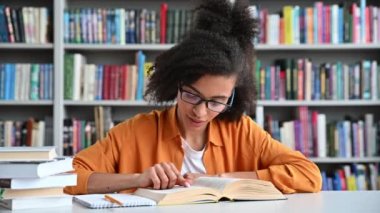 Zeki Afro-Amerikan öğrenci, şık kıyafetler ve gözlükler içinde, masada, kampüs kütüphanesinde, ilginç bir şekilde kitap okuyor, ders vermeye ya da sınava hazırlanıyor, bilgi ve öğrenim kavramı.