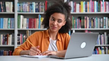 Kıvırcık saçlı, modaya uygun giyinmiş mutlu Afrikalı Amerikalı kız öğrenci, üniversite kütüphanesinde bir masada dizüstü bilgisayarla oturmuş, çevrimiçi dersler sırasında video çağrısından notlar alıyor, gülümsüyor.