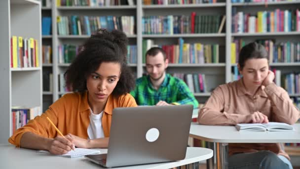 Studenten in der Vorlesung. Konzentrierte, ernsthafte afrikanisch-amerikanische Studentin sitzt am Tisch in der Universitätsbibliothek, bereitet sich auf Unterricht oder Prüfung vor, macht Notizen, benutzt Laptop, defokussierte Studenten im Hintergrund — Stockvideo