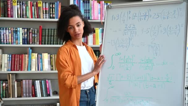 Modern African American smart young adult female teacher stand near the whiteboard, conducts online lecture by a video conference, looks at the webcam, shows information to the students, smiles — Stock Video