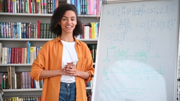 Αυτοπεποίθηση Αφρο-Αμερικανίδα έξυπνη νεαρή ενήλικη δασκάλα στέκεται κοντά στον πίνακα, διεξάγει διαλέξεις στη βιβλιοθήκη, δείχνει πληροφορίες στους μαθητές. Απομακρυσμένη μάθηση, online συνέδριο — Αρχείο Βίντεο