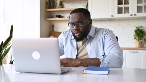 Удивленный афроамериканец с бородой, бизнес-директор, фрилансер, сидит за своим столом, смотрит на свой ноутбук с удивлением, снимая очки, читает неожиданные новости или плохие сообщения — стоковое видео
