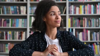 Güzel Afro-Amerikan kıvırcık saçlı kız, öğrenci ya da serbest çalışan, şık bir gömlek giyen, kütüphanede bir masada oturan, kameraya bakan, arkadaşça gülümseyen