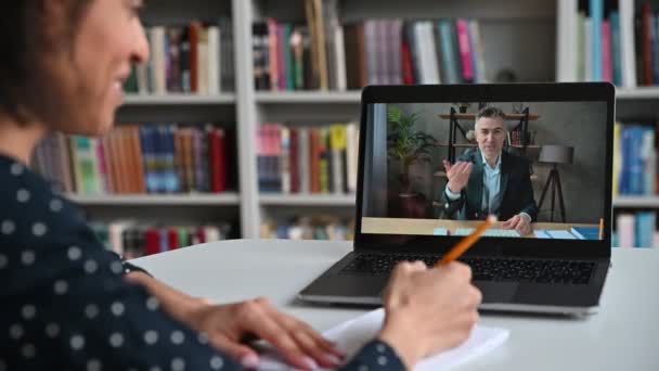 Vista por encima del hombro de una estudiante de raza mixta inteligente hasta la pantalla de un ordenador portátil con un maestro varón. Profesora madura lleva a cabo conferencia en línea, estudiante toma notas. Formación en línea, videollamada — Vídeos de Stock