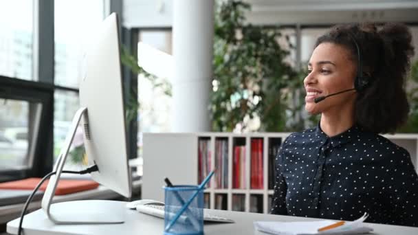 Φιλική γοητευτική νεαρή Αφροαμερικανή γυναίκα με ακουστικά, επιτυχημένη διευθύντρια ή τηλεφωνήτρια, κάθεται στο γραφείο, χρησιμοποιεί υπολογιστή, μιλώντας σε τηλεδιάσκεψη με πελάτη ή υπάλληλο, χαιρετώντας το χέρι — Αρχείο Βίντεο
