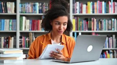 Mutlu akıllı konsantre olmuş Afrikalı Amerikalı öğrenci, not defterine notlar alıyor, online dersler dinliyor, bilgi kazanıyor, üniversite kütüphanesinde masada oturuyor, gülümsüyor.