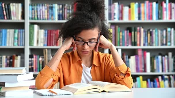 Moe uitgeput Afro-Amerikaanse studente zit aan een tafel met boeken in de bibliotheek, sluit haar ogen, masseert de brug van haar neus, hoofdpijn en stress van overwerk van het leren, moet rusten — Stockvideo