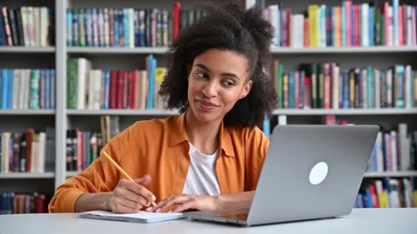 Happy african amerikansk smart kvinnelig student med krøllete hår, trendy kledd, sitter ved et skrivebord på universitetsbiblioteket, med en bærbar pc, tar notater i netttimen via videosamtale, smiler – stockvideo