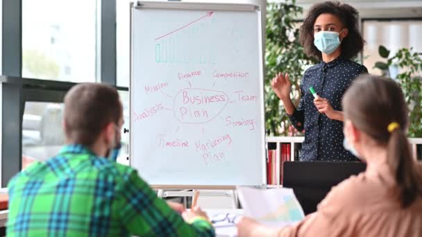 Negócios inteligente jovem afro-americano mulher, vestindo máscara médica protetora fica perto whiteboard no escritório, elegantemente vestida, mostra e explica plano de negócios e estratégia para seus colegas — Vídeo de Stock