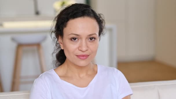 Portrait vidéo d'une charmante femme adulte métissée, portant un t-shirt blanc, assise à la maison sur le canapé, regardant directement la caméra avec un sourire agréable et convivial — Video