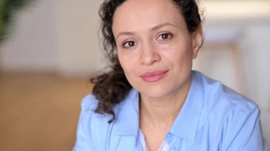 Çekici, kendine güvenen yetişkin, melez, esmer bir kadın, iş kadını, mavi bir gömlek giymiş, doğrudan kameraya bakıyor, dostça gülümsüyor.