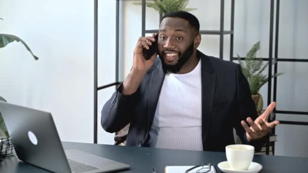 Удовлетворенный успешный афроамериканец, исполнительный или топ-менеджер, сидит на рабочем месте с, имеет приятный телефонный разговор с другом или коллегой. Телефонный разговор с использованием беспроводного смартфона — стоковое видео
