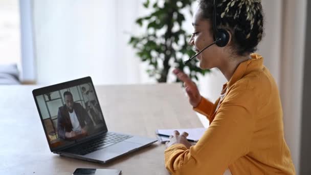 Vriendelijke jonge aantrekkelijke Afro-Amerikaanse vrouw in headset, call center werknemer, support operator, werken op een laptop, voert online overleg met de klant of collega, glimlachen. Videogesprek — Stockvideo