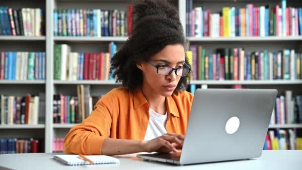 Chateada menina estudante afro-americana chocada, usando óculos, senta-se em uma mesa em uma biblioteca, usa laptop, apertou a cabeça em horror, olhando para a tela do laptop com expressão facial emocional estressada — Vídeo de Stock