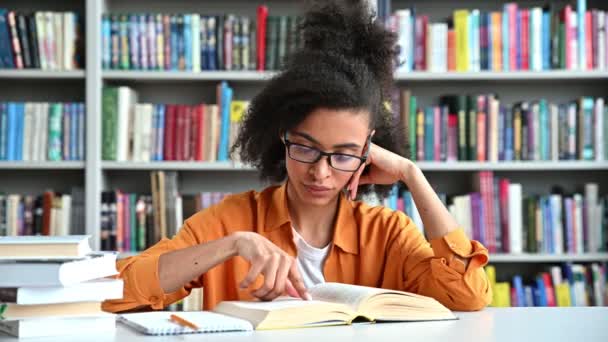 Egy okos, fókuszált afro-amerikai Y-generációs női hallgató ül egy főiskolai könyvtárban az asztalnál, érdeklődéssel olvas könyvet, felkészül az előadásra vagy vizsgára, fáradt és ásít, tudás és tanulás koncepció — Stock videók