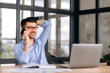Smartphone conversation. Satisfied friendly caucasian man, office worker or entrepreneur, sitting at a table in the office, having a pleasant phone conversation while working, smiling